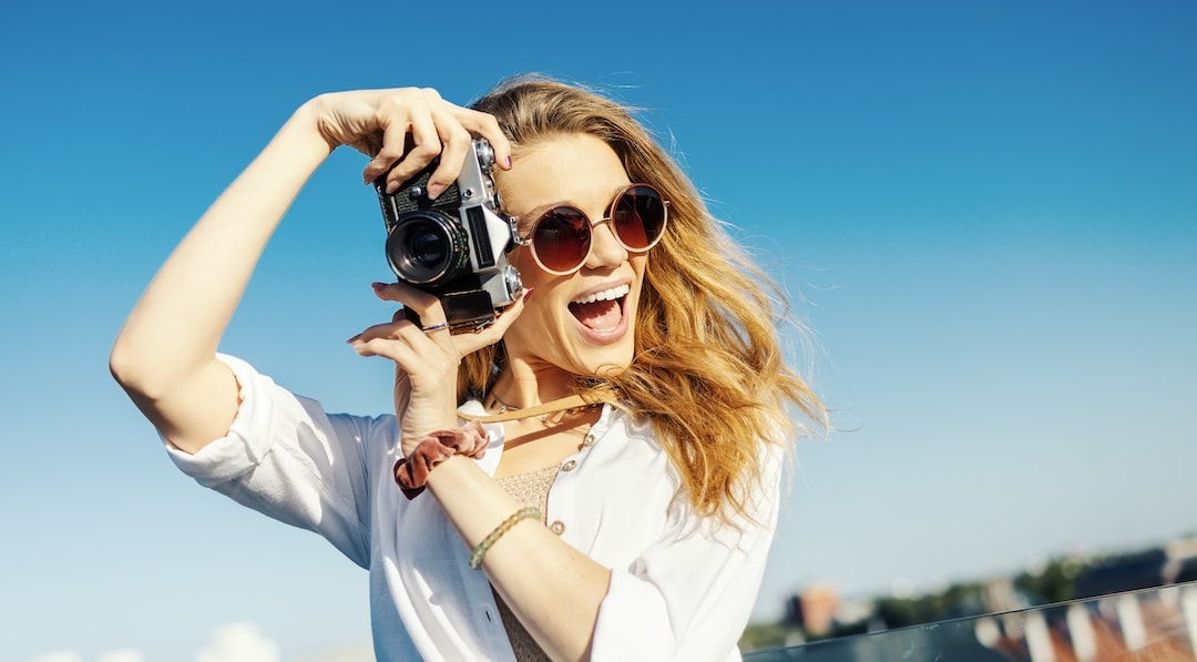 Close up shot of a smiling, fashionably dressed blonde woman posing with a camera